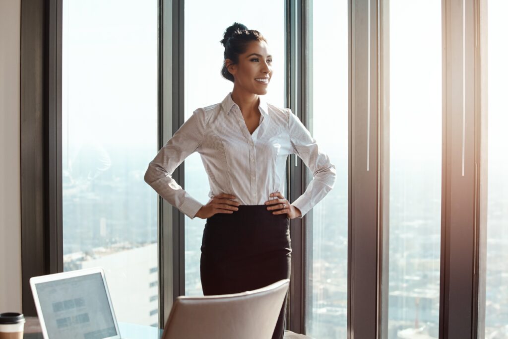 Confident Businesswoman Standing by Office Windows with Hands on Hips Embodying the Focus and Confidence of a High Performance Mindset