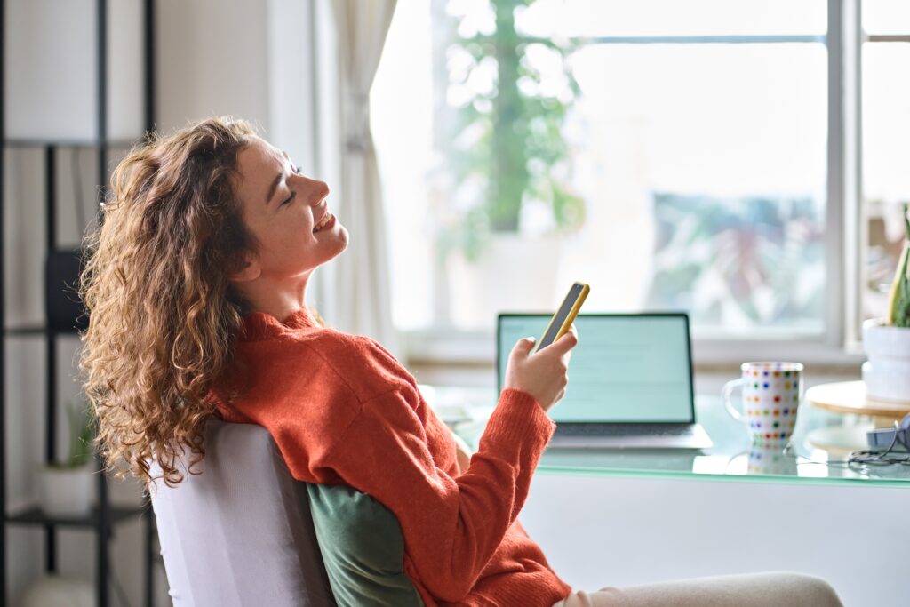 Relaxed Woman Smiling While Holding Her Phone at Her Desk Showing Balance and Confidencekey Qualities of What is a High Performer