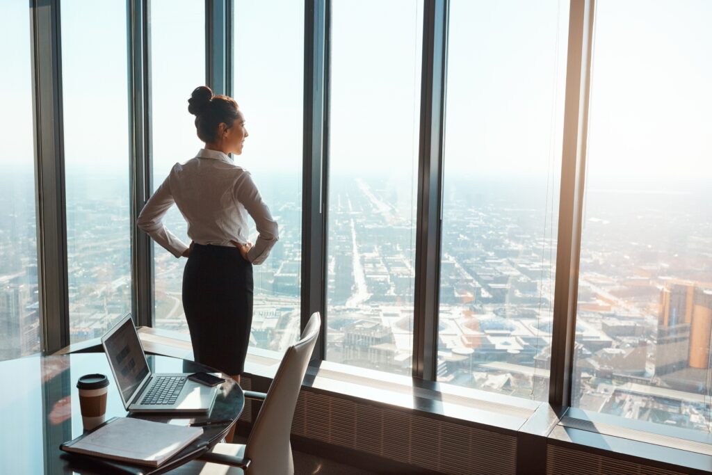 Confident Businesswoman Standing by Office Windows Overlooking the City Reflecting on Growth and Successshowing What It Means to Be a High Performer