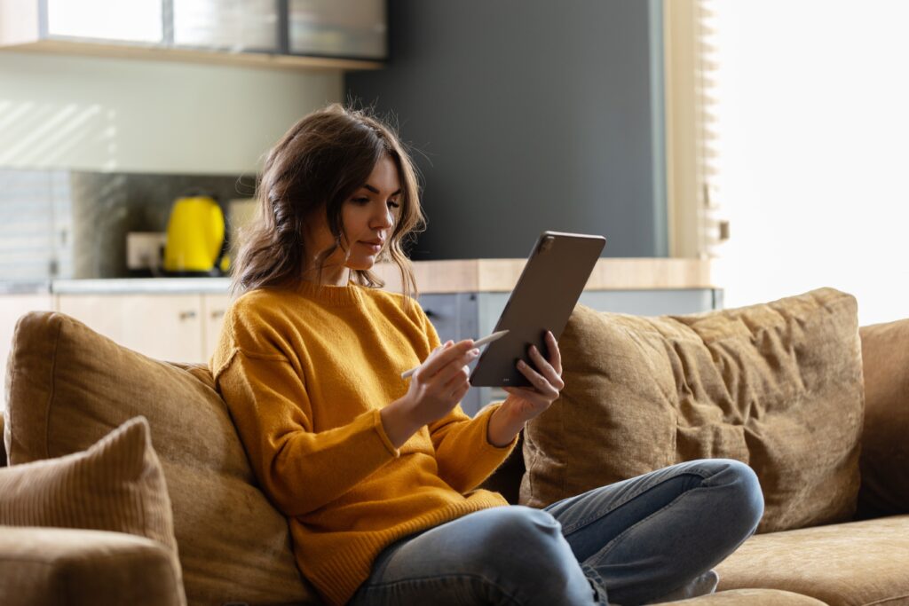 Woman Sitting on a Couch Using a Tablet Studying Through the High Performance Institute to Develop Advanced Coaching and Leadership Skills