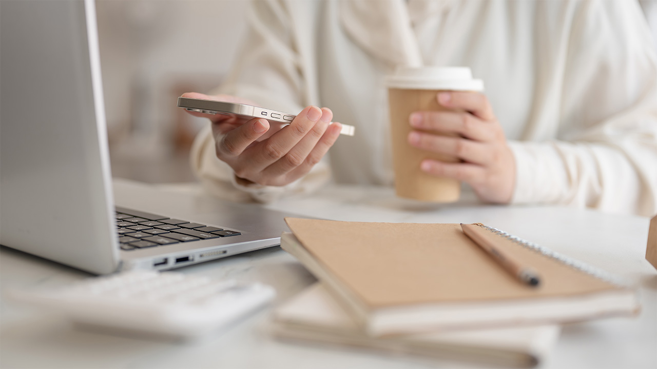 Close-up of a person holding a coffee and smartphone while working on a laptop, symbolizing focus, reflection, and the early signs you’re ready for high performance coaching.
