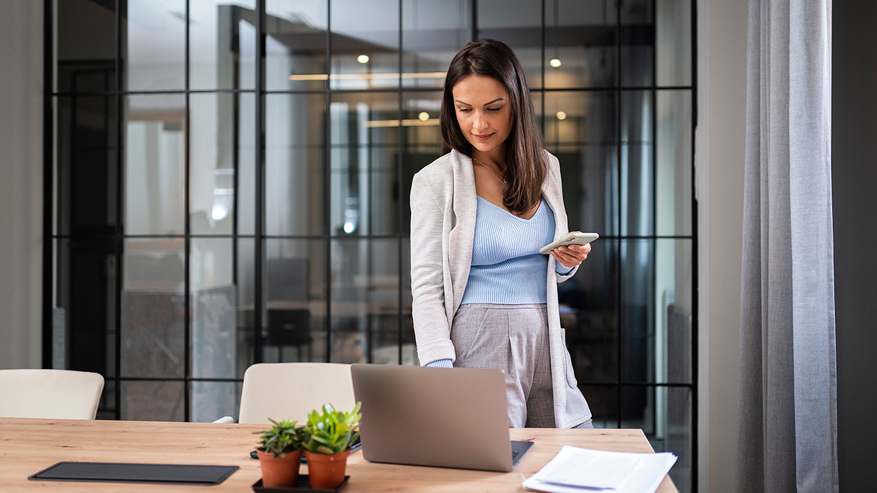 Focused businesswoman reviewing notes and using her phone beside a laptop, symbolizing decision-making and the research process for avoiding common mistakes when choosing a high performance coach.